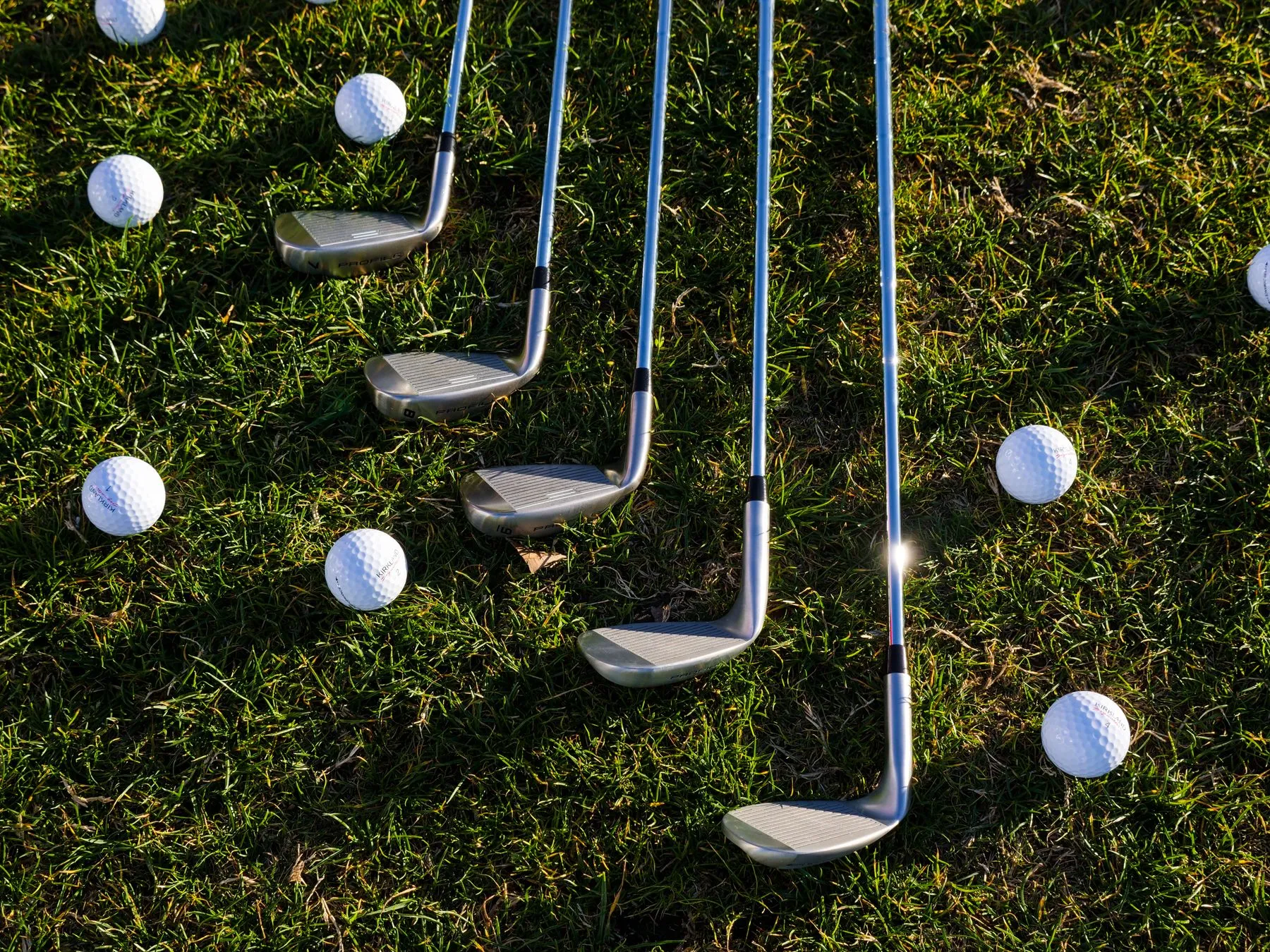 Row of steel golf irons and white golf balls laid out on sunlit grass
